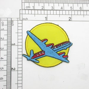 Può includere: Toppa ricamata raffigurante un aereo blu con dettagli rossi, su un cerchio giallo con bordo giallo. Nell'immagine è visibile un righello con misure in pollici e cm.