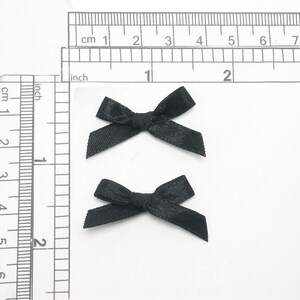 May include: Two black satin ribbon bows, each measuring approximately 1 inch in width, are displayed on a white surface. A ruler is visible in the background for size reference.