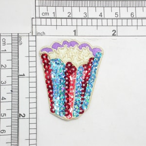 May include: A decorative sequin applique in a fan shape. The applique features red, blue, and iridescent sequins, with a cream-colored base and purple trim. The applique measures approximately 2 inches in height.