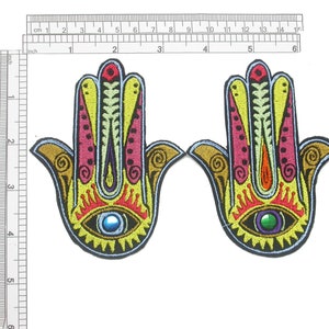 May include: Two embroidered Hamsa hand patches. Each patch features a detailed design with yellow, pink, and orange accents, a blue eye, and a gold border. The patches are approximately 7.6 cm tall.