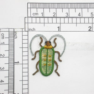 Könnte beinhalten: Grüne und gelbe gestickte Insekten-Applikation mit braunen Beinen und weißen Antennen. Die Applikation ist etwa 2,5 cm groß.