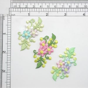 May include: Three embroidered flower appliques in pink, blue, and green. Each applique features a cluster of flowers with yellow centers and green leaves.