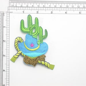 May include: A blue cowboy hat with a pink star on the front, surrounded by a green cactus and a yellow rope. The hat is embroidered with gold glitter.