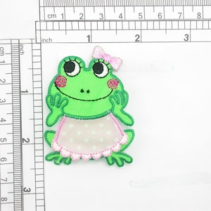 May include: A green frog patch with a pink bow, rosy cheeks, and a polka-dot apron. The embroidered patch measures approximately 2.5 inches in height and is designed for sewing or ironing onto fabric.