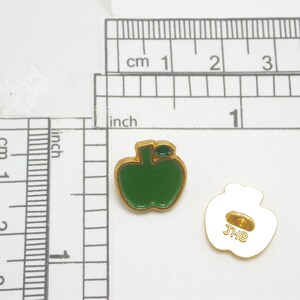 May include: Two apple-shaped pins. Each pin has a green enamel apple design with a gold-colored outline. The back of one pin is visible, showing a gold-colored clasp and the letters "JHB".