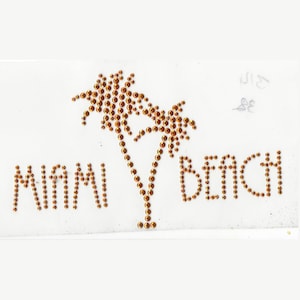Peut inclure: Un écusson en tissu blanc avec les mots "MIAMI BEACH" en strass dorés et un motif de palmier. L'écusson est probablement destiné aux vêtements ou aux accessoires. Le design est fait de petits strass ronds dorés.