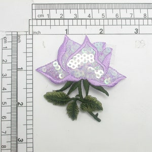 May include: A purple and white embroidered rose with sequins, approximately 6 cm in diameter, with green leaves and a stem.