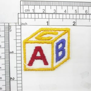 May include: A yellow, white, and blue embroidered patch shaped like a block with the letters A, B, and C on each side.