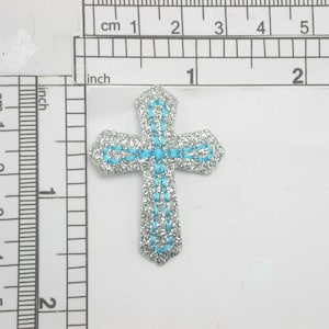 Może przedstawiać: Srebrny krzyż z brokatem z niebieskim haftowanym konturem. Krzyż ma około 2,5 cm.