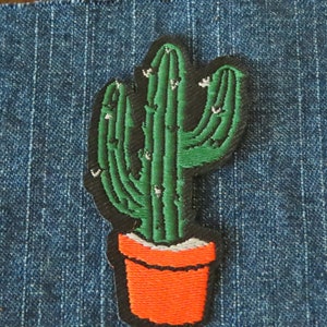 Pode incluir: Um cacto verde com detalhes brancos em um vaso vermelho. O cacto é bordado em um tecido jeans azul.