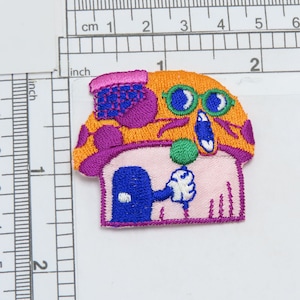 May include: A colourful embroidered patch featuring a cartoon character with a pink and orange hat, blue eyes, and a green ball. The character is holding a white object in its hand. The patch is outlined in purple.
