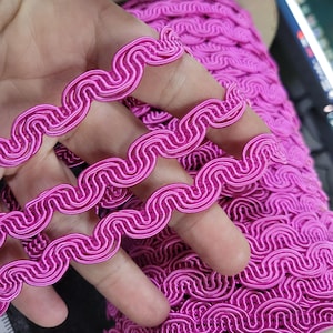 Op de afbeelding: Close-up van een rol felroze decoratieve bies met een golvend, gelust ontwerp. De bies is gemaakt van een glanzend, geweven materiaal. De bies wordt in een hand gehouden.