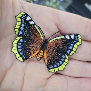May include: Embroidered butterfly patch. The butterfly has a brown body and wings with yellow, blue, and white details. The patch is intricately stitched, giving a textured look. A decorative item for clothing or accessories.