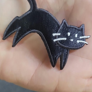May include: An embroidered fabric patch shaped like a black cat in a leaping or stretching pose. The cat has a solid black body with stitched white thread details for the whiskers and facial features, featuring a curved tail and pointed ears.