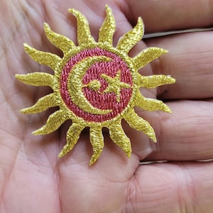 Peut inclure: Un écusson brodé circulaire représentant un soleil doré métallique avec des rayons pointus rayonnants entourant un centre rose foncé. Le centre contient un croissant de lune doré et une petite étoile à cinq branches en fil.
