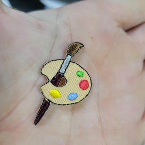 Pode incluir: Remendo bordado com uma paleta de artista e um pincel. A paleta é bege com manchas de tinta amarela, verde, vermelha e azul. O pincel tem uma escova marrom e um cabo vermelho escuro.