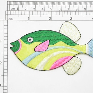 May include: An embroidered iron-on patch of a cartoon fish, featuring green, yellow, pink and blue colours. The fish has a large, round body and a small, pointed tail.