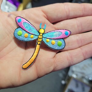 May include: A colorful dragonfly patch with embroidered details. The wings are light blue with pink edges and dotted with yellow and green circles. The body is yellow and the edges are black.