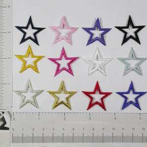 Puede incluir: Doce parches termoadhesivos en forma de estrellas con un contorno blanco. Las estrellas son de varios colores, incluyendo negro, rosa, morado, dorado, plateado, rojo y azul.