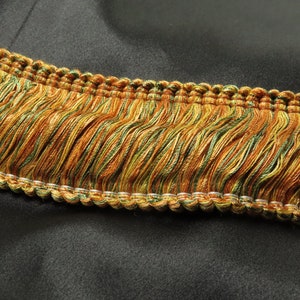 May include: A close-up of a gold and green fringe trim with a woven edge. The fringe is made of a soft, silky material and has a subtle sheen.