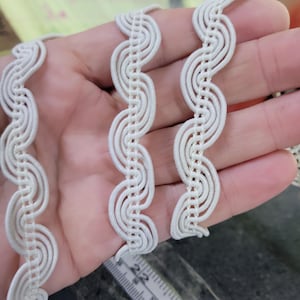 May include: Off-white decorative trim with a wavy design. The trim is made of a woven material and is held in a hand. The trim is measured with a ruler.