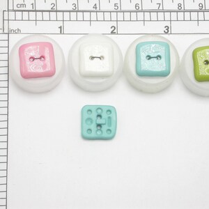May include: A collection of colorful square buttons in pink, white, turquoise, and green, each with two holes for sewing. The buttons are set on a white background with a ruler for scale. A smaller turquoise button is also present.