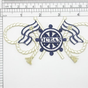 May include: An embroidered patch in blue and white with a nautical theme. It features a ship's wheel with the word "OCEAN" in the centre, two crossed flags with stars, and gold rope accents.