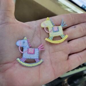 May include: Two embroidered rocking horse patches. One is light blue with pink and yellow accents, the other is yellow with blue and pink accents. Both have a saddle and are on a light background.
