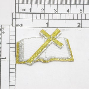 Op de afbeelding: Een witte geborduurde patch met een gouden kruis en een boek. Het kruis bevindt zich bovenop het boek.
