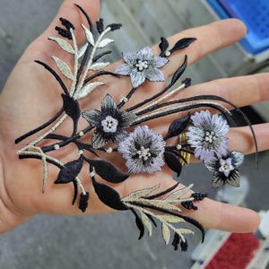 May include: Embroidered floral applique with black, silver, and lavender flowers and leaves. The design has detailed stitching and a delicate, ornamental appearance. Suitable for embellishing garments or craft projects.