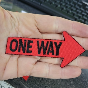 May include: A red arrow-shaped patch with the words "ONE WAY" embroidered in black. The patch has a black border and is likely designed to be sewn onto clothing or accessories. The arrow points to the right.