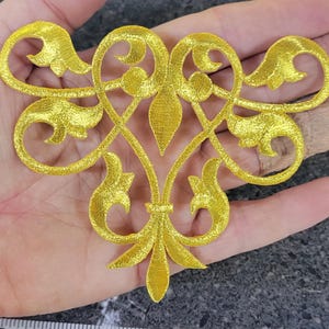 May include: A bright yellow embroidered appliqué featuring a symmetrical scrollwork design with curved lines and pointed leaf-like motifs. A white measuring tape is positioned underneath, showing the piece spans approximately 10 cm in width.
