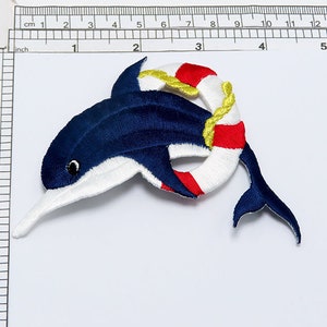 May include: Embroidered iron-on patch of a blue dolphin wearing a red and white life preserver with a yellow rope.