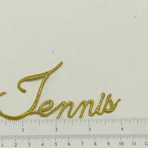 May include: Gold glitter iron-on patch with the word "Tennis" in a cursive font.
