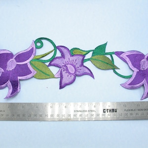 以下が含まれることがあります： 緑の葉と蔓のある紫色の刺繍された花のトリム。トリムの長さは約33cmです。