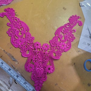 May include: A vibrant fuchsia lace applique with an intricate floral and leaf design. The applique is a V-neck shape, perfect for embellishing garments or craft projects. A ruler with inch measurements is visible.