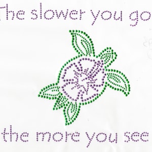 Può includere: Una tartaruga verde e viola fatta di strass con il testo "The slower you go... the more you see" in strass viola.