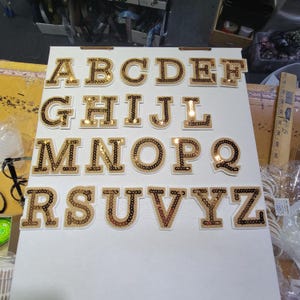 May include: A complete alphabet of gold sequined letters, arranged on a white surface. Each letter is individually cut with a lighter gold outline and sequined fill. The letters are in a serif font.