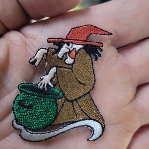May include: An embroidered patch featuring a witch with black hair, a red pointed hat, and a brown robe, stirring a bright green cauldron. The witch has pale skin and is depicted in a side profile view with a white curved base element underneath.
