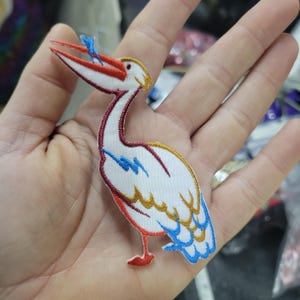 May include: Embroidered patch featuring a white bird with a long red and white beak, holding a blue fish. The bird has gold, blue, and red accents. The patch is likely for clothing or accessories.