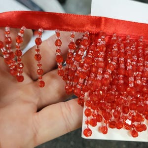 May include: Red beaded fringe trim with a satin ribbon backing. The fringe is made of small, faceted red beads, creating a shimmering effect. Ideal for embellishing clothing, accessories, or home decor projects.