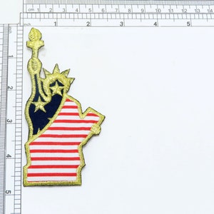 May include: Gold embroidered patch of the Statue of Liberty with red and white stripes, resembling the American flag.