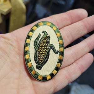 May include: Oval embroidered patch featuring a detailed corn on the cob design. The corn is yellow with green husks, set against a cream background. The border is a pattern of green and gold squares.