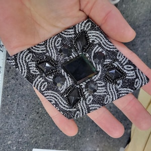 May include: A black and silver decorative patch with a diamond shape. The patch features a swirling silver pattern on a black background, with black gemstones and beads. The patch is held in a hand.