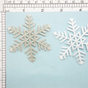 May include: Two snowflake-shaped fabric patches, one silver and one white, on a light blue background. The silver snowflake has a glittery finish. The white snowflake is plain. Both snowflakes are approximately 2 inches in diameter.