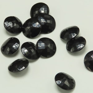 May include: A group of black, faceted, oval beads. The beads have a glossy finish and reflect light, creating a sparkling effect. They are scattered on a white surface, suggesting a craft or jewellery-making theme.