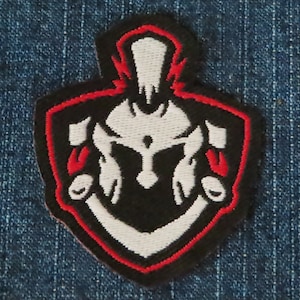 May include: A black and white embroidered patch with a red outline. The patch features a stylised image of a warrior's helmet.