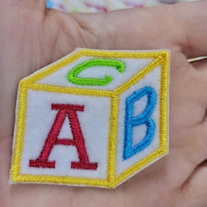 May include: A white fabric patch shaped like a cube with yellow embroidered borders. The front face features a red letter A, the right side features a blue letter B, and the top face features a green letter C, all rendered in embroidery.
