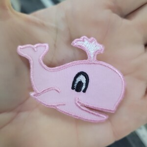 May include: A pink embroidered whale patch with a black eye and white water spout. The patch has a light pink background and a pink outline. Suitable for clothing, bags, or other fabric items.