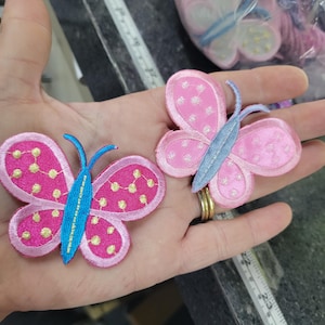May include: Two pink butterfly patches with blue accents are displayed. The butterfly wings have a dotted pattern in yellow. The butterflies are embroidered and have a light blue body and antennae.
